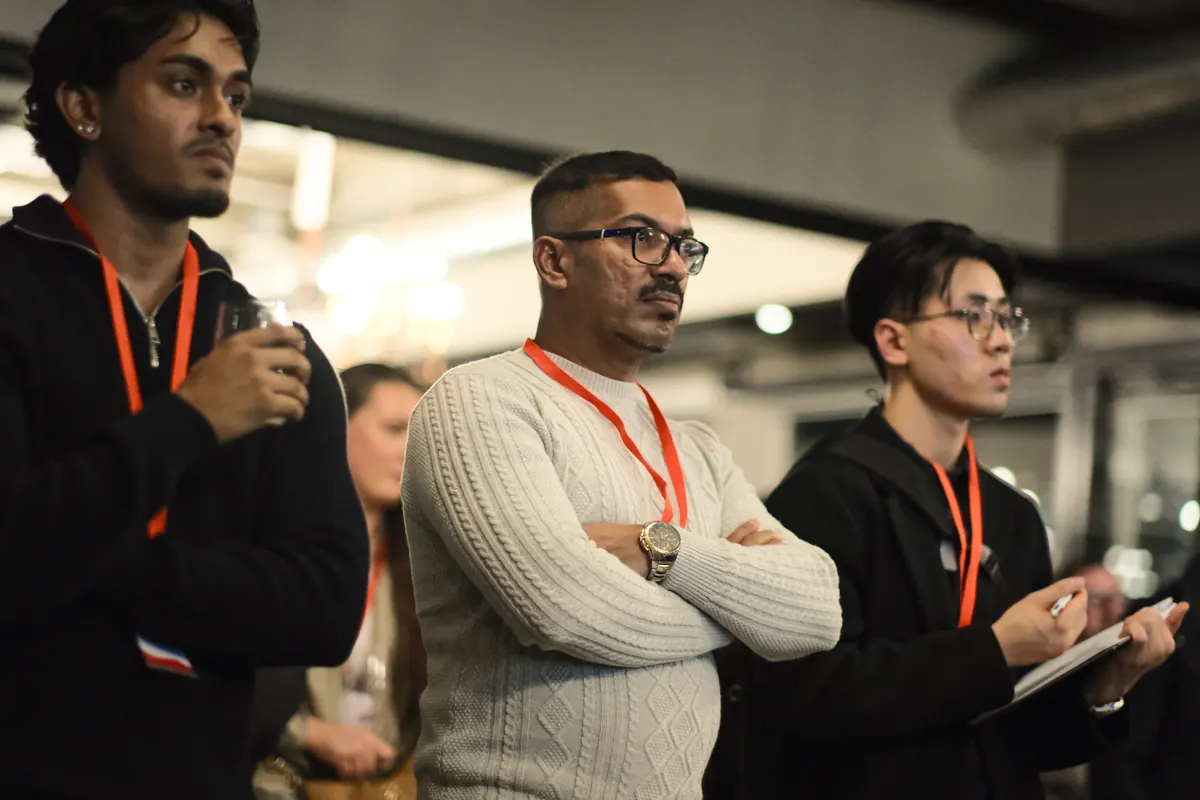 Three members standing and listening at a BNB Success event; the one on the right writes notes on a pad.