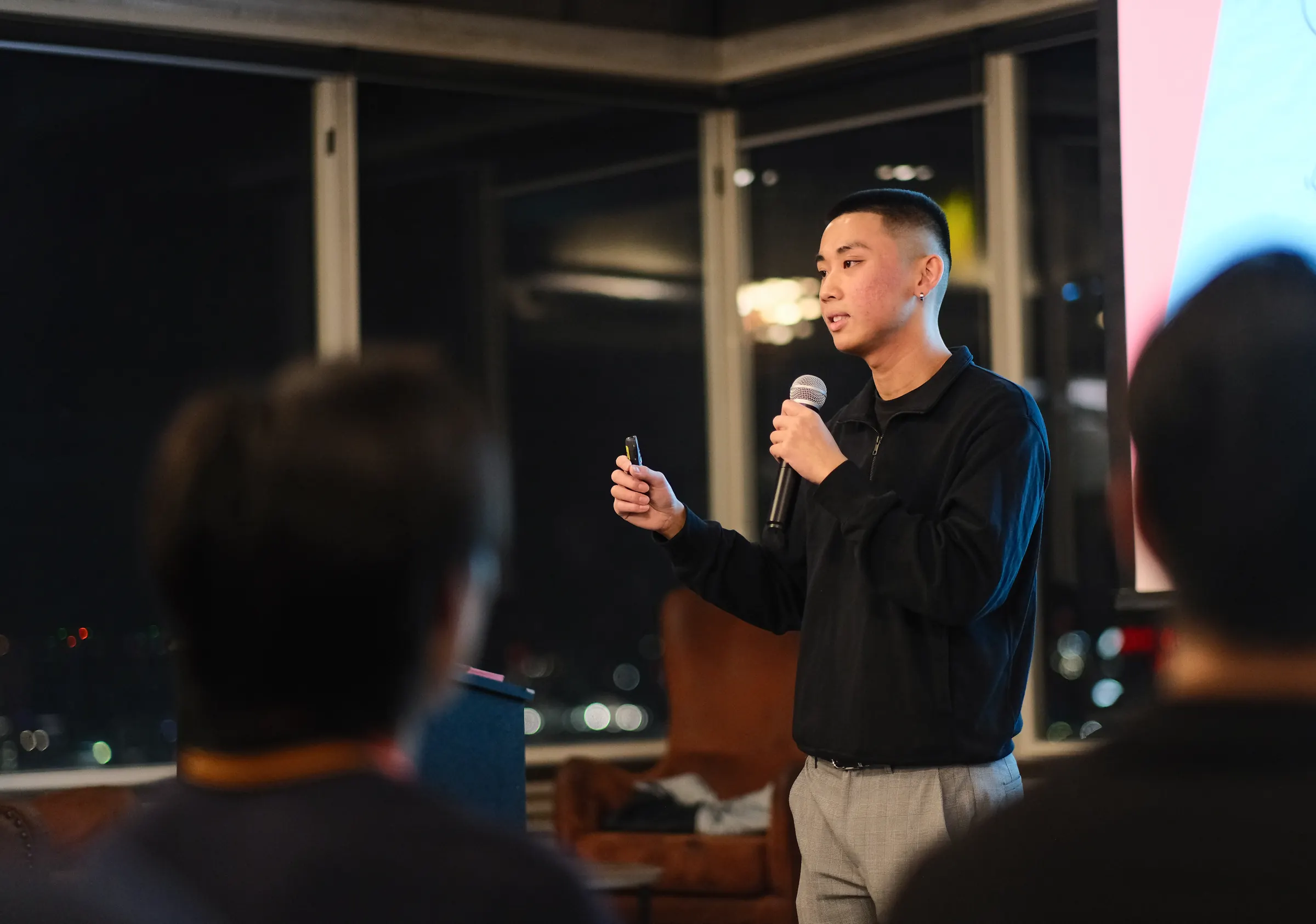 Jordan Pham speaking with a microphone at a BNB Success event, seen over the shoulders of the audience, with city lights in the windows behind him.