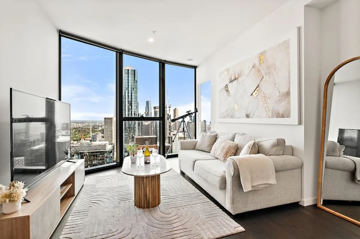 A compact living room with a beige sofa, a round timber coffee table and a window wall over the city.