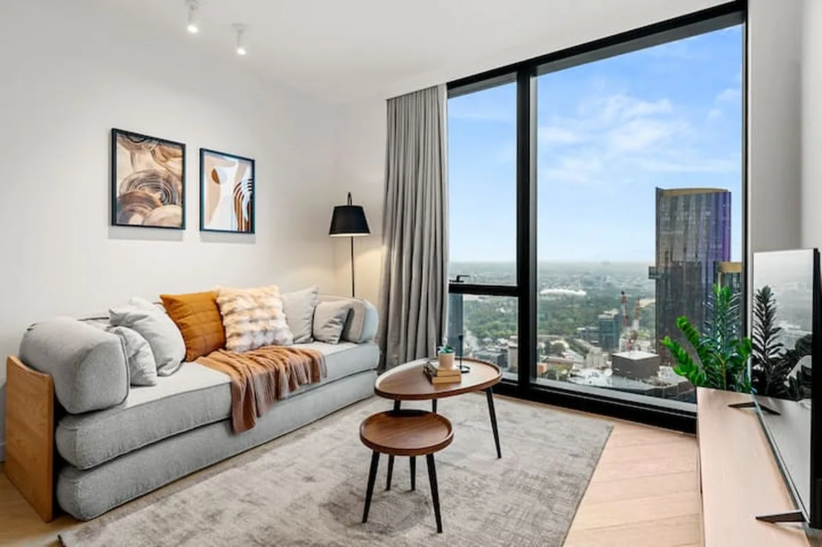 A furnished high-rise living room with a grey sofa, orange cushions and floor-to-ceiling windows over Melbourne.