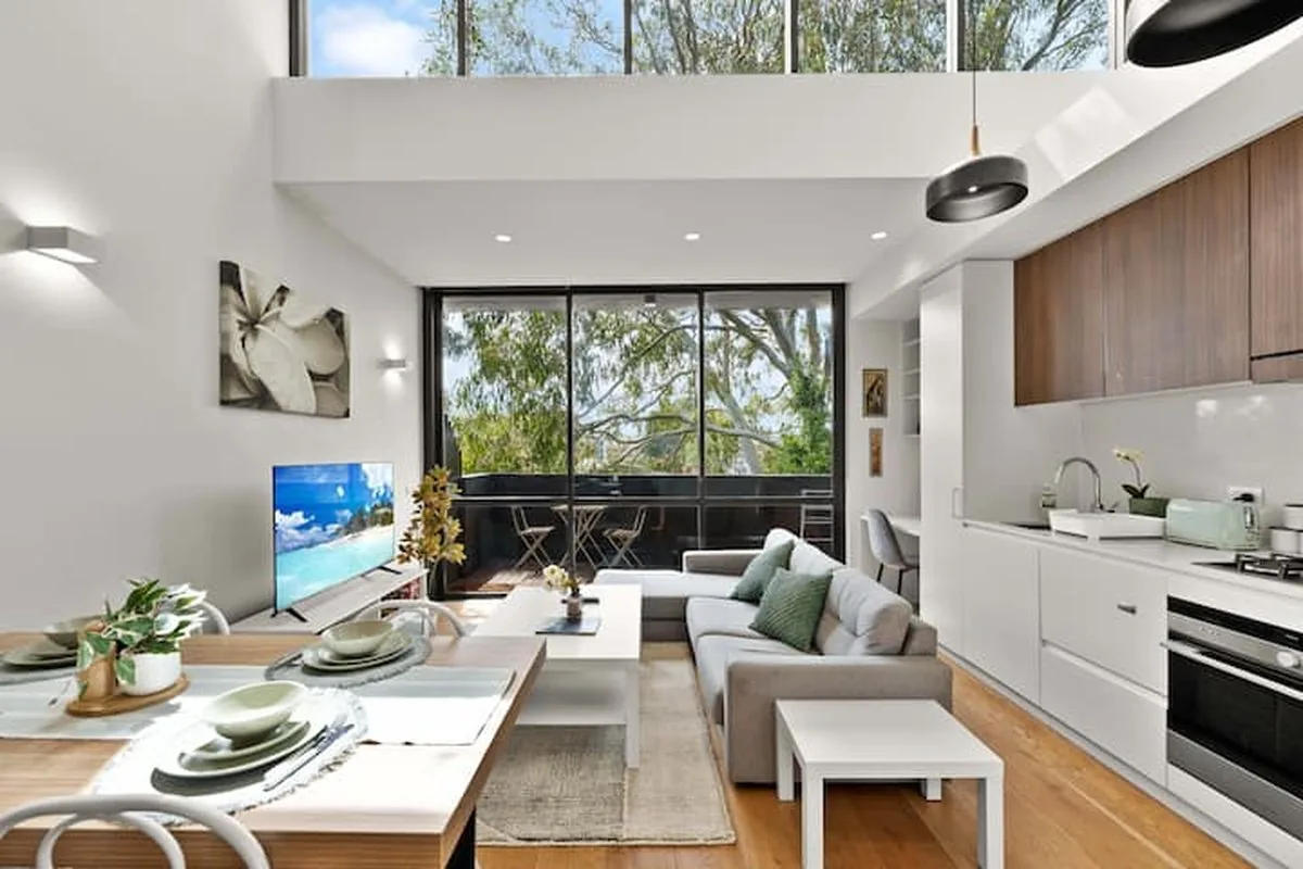 A double-height open-plan living and kitchen area with a timber dining table set for four and trees outside the balcony doors.
