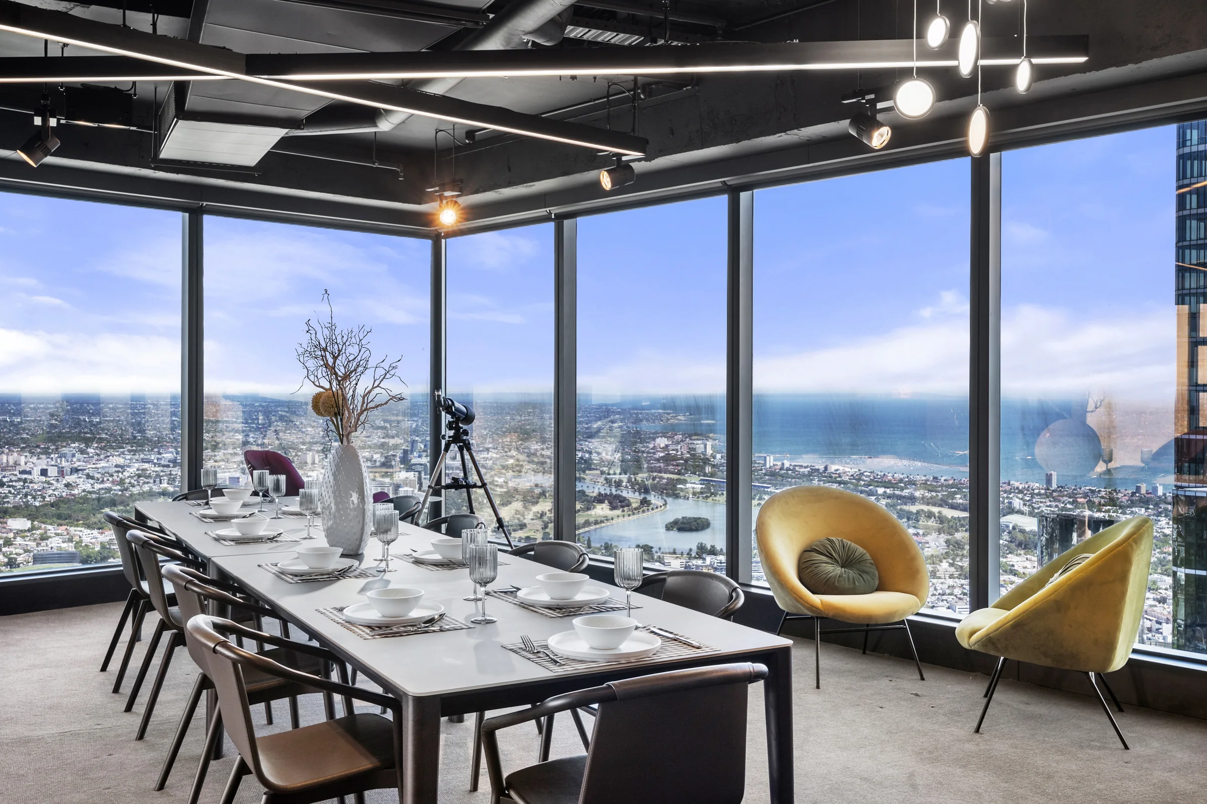 A dining table laid for six in a top-floor apartment, with floor-to-ceiling windows looking over the city and the bay.